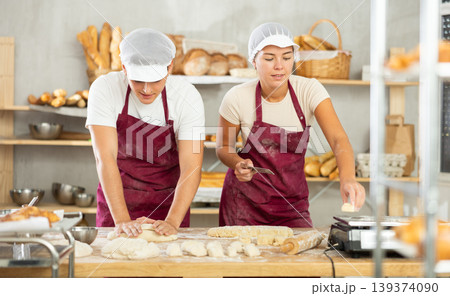 Two young bakers prepare dough in kitchen of bakery - together they knead dough, cut it with knife and weigh it on scales 139374090
