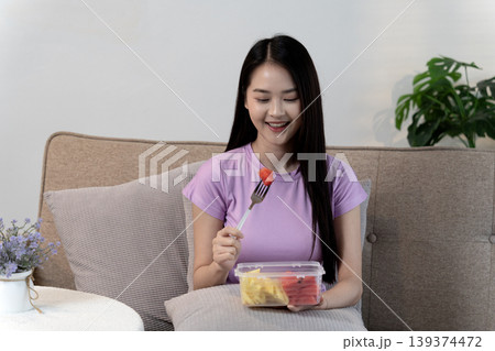 Asian young adult woman eating healthy fruit salad on sofa at home for wellness and healthy lifestyle Asian young adult woman eating healthy fruit salad on sofa at home for wellness and healthy lifestyle 139374472