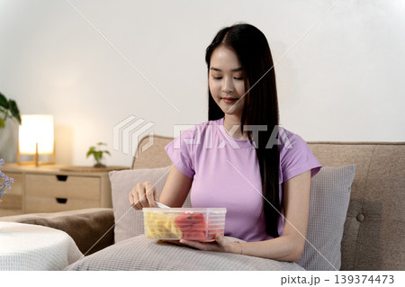 Asian woman adult enjoy healthy fruit snack home sofa eating fresh watermelon pineapple living room wellness lifestyle self care for balanced diet 139374473