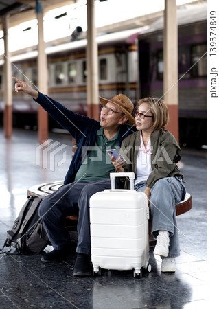 Asian adult couple travelers discuss train journey tourism destination pointing smartphone at railway station platform enjoying vacation leisure trip Asian adult couple travelers discuss train journey tourism destination pointing smartphone at railway station platform enjoying vacation leisure trip 139374849