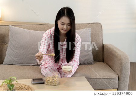 Asian young adult woman eats healthy breakfast cereal with yogurt at home living room morning relaxation cozy comfortable lifestyle Asian young adult woman eats healthy breakfast cereal with yogurt at home living room morning relaxation cozy comfortable lifestyle 139374900