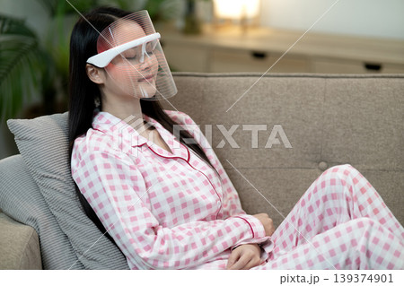 Asian adult woman applying LED light therapy mask home for skin care beauty treatment facial rejuvenation anti aging relaxation living room couch Asian adult woman applying LED light therapy mask home for skin care beauty treatment facial rejuvenation anti aging relaxation living room couch 139374901