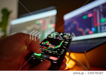 Trader using smartphone with stock market chart and monitors Trader using smartphone with stock market chart and monitors 139376387