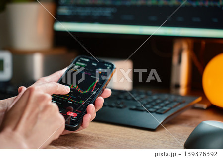 Person using trading app on smartphone at desk with market chart on screen. Person using trading app on smartphone at desk with market chart on screen. 139376392