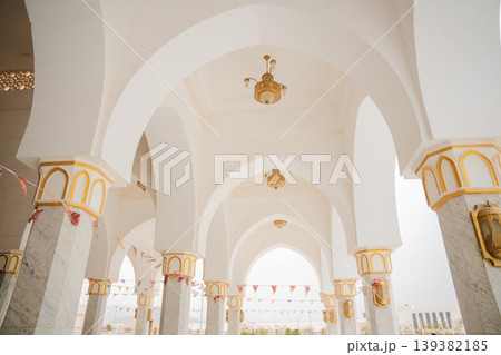 Tall white arches and marble columns with gold accents in a mosque courtyard with hanging lanterns and colorful flags. Islamic religion, architecture, exterior space Tall white arches and marble columns with gold accents in a mosque courtyard with hanging lanterns and colorful flags. Islamic religion, architecture, exterior space 139382185