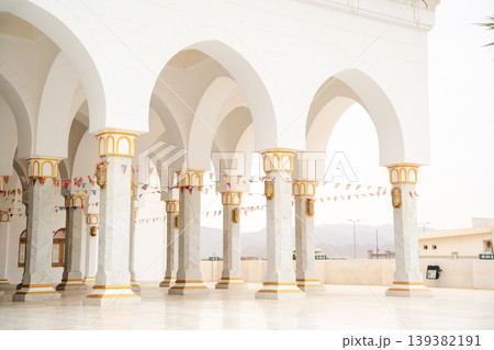 Mosque colonnade with white arches and marble columns decorated with gold details, open courtyard with hanging lanterns and flags visible. Islamic architecture and cultural heritage in an arid region Mosque colonnade with white arches and marble columns decorated with gold details, open courtyard with hanging lanterns and flags visible. Islamic architecture and cultural heritage in an arid region 139382191