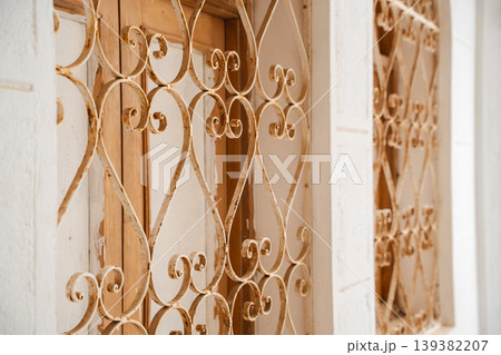 Close up of mosque window wrought iron grille with curved scroll pattern mounted over wooden shutters in white wall frame. Islamic architecture, decorative metalwork, exterior detail Close up of mosque window wrought iron grille with curved scroll pattern mounted over wooden shutters in white wall frame. Islamic architecture, decorative metalwork, exterior detail 139382207