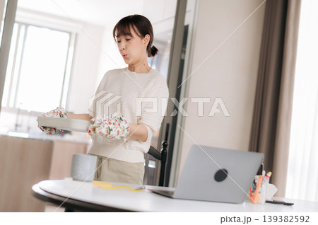 Young asian woman stands near table holding floral arrangement looking focused towards laptop computer screen in bright contemporary apartment living room space 139382592