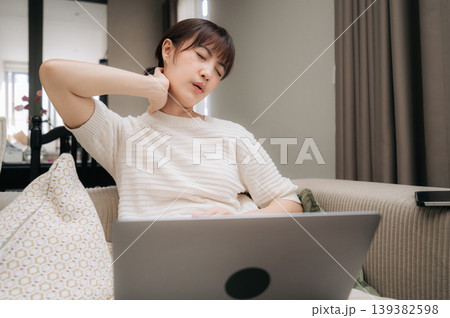 Exhausted young asian woman feeling severe neck pain while working on laptop sitting on cozy sofa at home showing deeply tired and stressed emotional facial expression 139382598