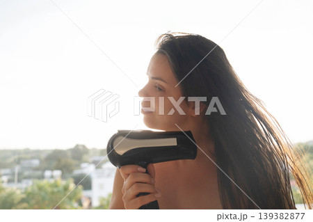 Female model drying hair with hairdryer near window view, beauty routine with natural sunlight, attractive woman with healthy skin and long hair, wellness and lifestyle concept 139382877