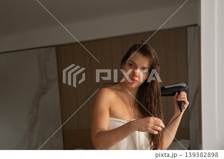 Attractive woman drying long wet hair with hair dryer in front of mirror healthy shiny hair beauty routine indoor portrait Attractive woman drying long wet hair with hair dryer in front of mirror healthy shiny hair beauty routine indoor portrait 139382898