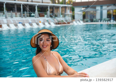 Attractive woman leaning on pool edge smiling in straw hat and bikini enjoying summer vacation resort lifestyle beauty portrait with turquoise water background 139382917