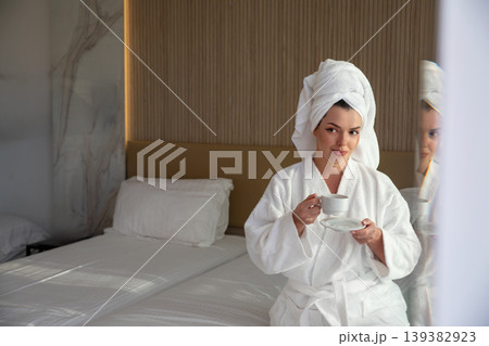 Beautiful woman in white bathrobe with towel on head holding coffee cup sitting on bed enjoying calm morning beauty lifestyle and relaxation in modern hotel room 139382923