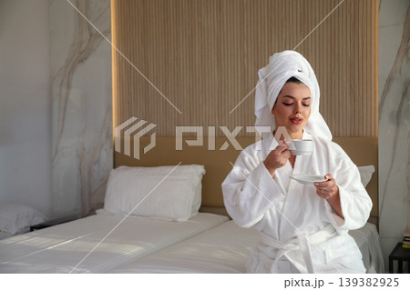Pretty woman in bathrobe and towel enjoying coffee in bed smiling softly in cozy hotel room morning light beauty lifestyle relaxation portrait Pretty woman in bathrobe and towel enjoying coffee in bed smiling softly in cozy hotel room morning light beauty lifestyle relaxation portrait 139382925