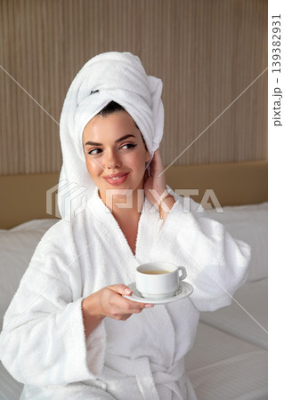 Beauty woman enjoying peaceful morning coffee in bathrobe sitting on bed smiling softly beauty skincare wellness lifestyle calm hotel interior relaxation concept 139382931