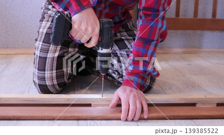 A worker in a plaid shirt operates a screwdriver, driving fasteners into the side panel of a light wood bed on the floor of a room. 139385522