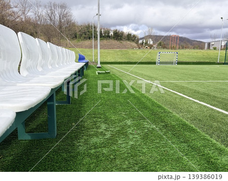 A soccer field, featuring a row of white plastic stadium seats with green metal frames running along the left edge. A soccer field, featuring a row of white plastic stadium seats with green metal frames running along the left edge. 139386019