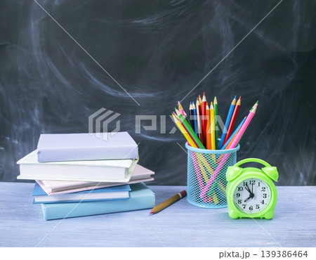School supplies with books, pencils and alarm clock on the table against a blackboard background, back to school concept. School supplies with books, pencils and alarm clock on the table against a blackboard background, back to school concept. 139386464