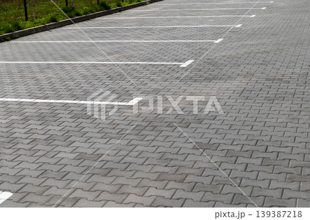 A parking lot shows empty spaces with white lines on gray paver stones under bright daylight. Grass is visible nearby A parking lot shows empty spaces with white lines on gray paver stones under bright daylight. Grass is visible nearby 139387218