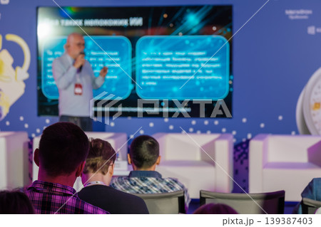 Business conference presentation with speaker on stage and attentive audience in chairs Business conference presentation with speaker on stage and attentive audience in chairs 139387403
