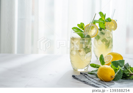 A summer drink with lemon, mint, and ice in a two glasses against a sunny window background. 139388254