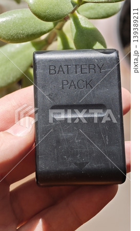Black battery pack held in a hand with green succulent plant in the background, showcasing the product's design and texture under natural light Black battery pack held in a hand with green succulent plant in the background, showcasing the product's design and texture under natural light 139389211