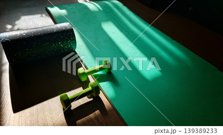 Green exercise mat with yellow dumbbells and a black foam roller positioned on a wooden floor, sunlight casting shadows across the scene Green exercise mat with yellow dumbbells and a black foam roller positioned on a wooden floor, sunlight casting shadows across the scene 139389233