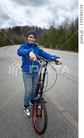 Young boy wearing blue jacket and gray beanie stands beside bicycle on empty road surrounded by trees under cloudy sky 139389239