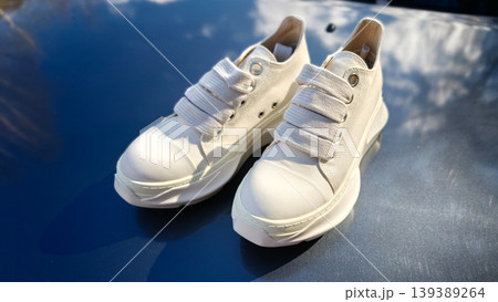 White casual sneakers with textured fabric and laces displayed on a reflective surface under natural sunlight, showcasing modern footwear design and style 139389264