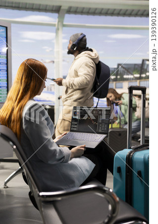 Independent female trader checking financial data and currency exchange, investment performance on laptop in airport waiting area. Busy terminal emphasizes remote trading and nomadic lifestyle. Independent female trader checking financial data and currency exchange, investment performance on laptop in airport waiting area. Busy terminal emphasizes remote trading and nomadic lifestyle. 139390126