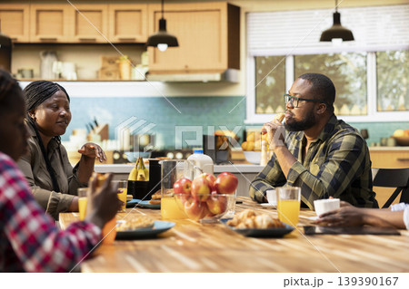 Family enjoying a weekend breakfast together in a sunny kitchen. Parents serve coffee, croissants and orange juice while children laugh, moment of love and happiness around the table. 139390167