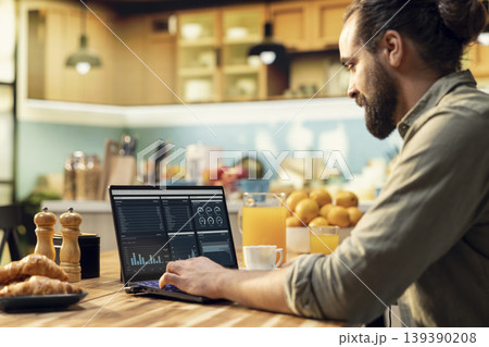 Remote worker using laptop and digital tools to analyze data and research information, seated in the kitchen. Telecommuting and outsourcing with charts and network connectivity, modern lifestyle. 139390208