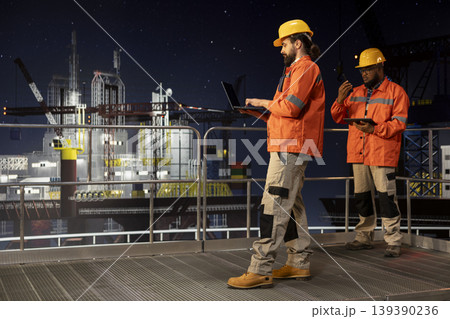 Barge engineering team on offshore platform deck monitoring drilling operations during night shift. Drilling rig coworkers reviewing sensor data on devices to ensure continuous production 139390236