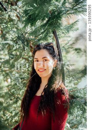 Young Caucasian woman with long dark hair wearing red sweater standing among green tree branches in park. Outdoor portrait and natural lifestyle theme Young Caucasian woman with long dark hair wearing red sweater standing among green tree branches in park. Outdoor portrait and natural lifestyle theme 139390295