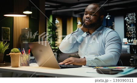 African american male worker stretching his back and neck muscles, correcting posture for muscle relief and relaxation. Strain and neck stiffness due to intense activity in office. African american male worker stretching his back and neck muscles, correcting posture for muscle relief and relaxation. Strain and neck stiffness due to intense activity in office. 139390311