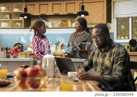 Busy african american working dad typing on laptop at table with his family, ignoring his small children and wife during breakfast. Distracted preoccupied father distant with his two kids. Busy african american working dad typing on laptop at table with his family, ignoring his small children and wife during breakfast. Distracted preoccupied father distant with his two kids. 139390349