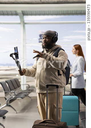 Happy black blogger filming vlog in the airport waiting area, sharing travel tips through smartphone. Modern online presence and the lifestyle of a solo traveler creating engaging content. Happy black blogger filming vlog in the airport waiting area, sharing travel tips through smartphone. Modern online presence and the lifestyle of a solo traveler creating engaging content. 139390473