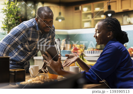 Smiling boyfriend and girlfriend sharing breakfast pastries and coffee at home. Laughing and bonding over casual conversation, watching funny videos on tablet for a candid cute moment. 139390513