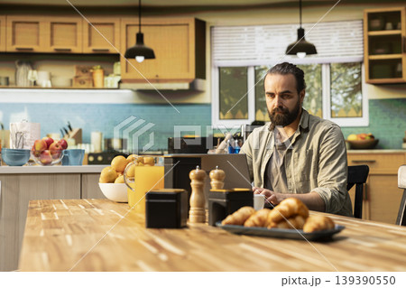 Adult man working from home in a cozy rustic kitchen, using laptop for daily tasks online. Freelancer lifestyle with remote work setup and natural light supporting productivity and multitasking. 139390550