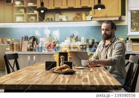 Adult man working from home in a cozy rustic kitchen, using laptop for daily tasks online. Freelancer lifestyle with remote work setup and natural light supporting productivity and multitasking. Adult man working from home in a cozy rustic kitchen, using laptop for daily tasks online. Freelancer lifestyle with remote work setup and natural light supporting productivity and multitasking. 139390556