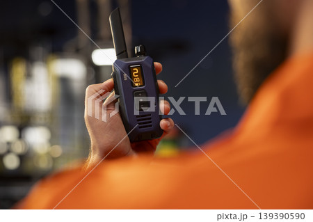 Closeup of offshore rig employee using walkie talkie to coordinate safety protocols with logistics team at night. Drilling barge specialist talks into handheld radio device, supervising deck equipment Closeup of offshore rig employee using walkie talkie to coordinate safety protocols with logistics team at night. Drilling barge specialist talks into handheld radio device, supervising deck equipment 139390590