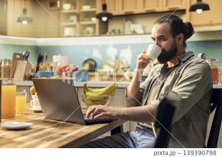 Adult man, morning coffee, working from home, kitchen environment, laptop for online work. Remote worker lifestyle, wireless internet and modern technology, multitasking, digital nomad. Adult man, morning coffee, working from home, kitchen environment, laptop for online work. Remote worker lifestyle, wireless internet and modern technology, multitasking, digital nomad. 139390798