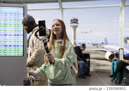 Influencer girl live streaming from airport lounge with phone and tripod, interacting with audience before flight departure. Content creator culture, digital nomad flexibility and travel vlog. Influencer girl live streaming from airport lounge with phone and tripod, interacting with audience before flight departure. Content creator culture, digital nomad flexibility and travel vlog. 139390901