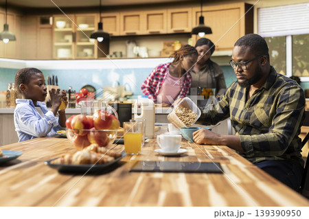 Cheerful black family enjoying breakfast together in a bright cozy kitchen. Parents serve toast and coffee while children laugh at the table, creating a warm morning filled with happiness. Cheerful black family enjoying breakfast together in a bright cozy kitchen. Parents serve toast and coffee while children laugh at the table, creating a warm morning filled with happiness. 139390950