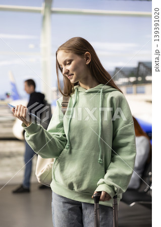 Gen z girl traveler in airport waiting area verifies information on plane ticket, busy terminal activity with baggage and passengers. Tourism, economy travel and real-life airport experiences. Gen z girl traveler in airport waiting area verifies information on plane ticket, busy terminal activity with baggage and passengers. Tourism, economy travel and real-life airport experiences. 139391020
