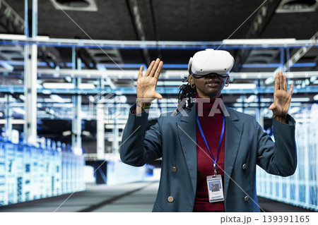Data center female programmer using VR headset gear to configure server settings. IT specialist in modern high tech server hub uses virtual reality technology to do maintenance tasks on infrastructure 139391165