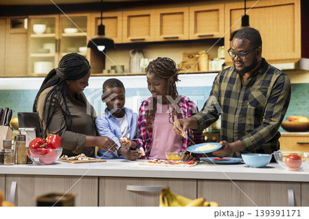 African American parents and kids sharing pizza day at home, adding mozzarella over the dough with multiple ingredients. Togetherness, laughter, family bonding and homemade cooking. African American parents and kids sharing pizza day at home, adding mozzarella over the dough with multiple ingredients. Togetherness, laughter, family bonding and homemade cooking. 139391171
