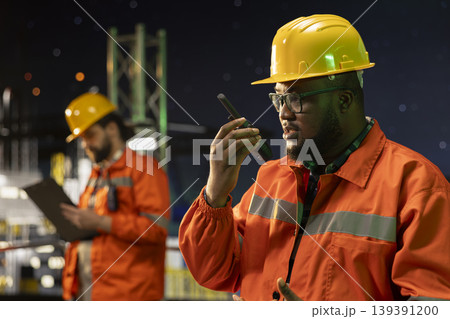 Close up of drilling barge technician coordinating remote team tasks using portable radio system. African american man speaks into walkie talkie mic, manage logistics on maritime offshore platform Close up of drilling barge technician coordinating remote team tasks using portable radio system. African american man speaks into walkie talkie mic, manage logistics on maritime offshore platform 139391200
