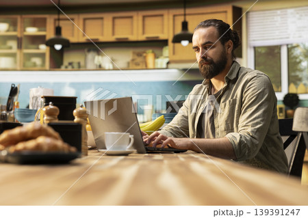 Remote worker, kitchen table with laptop, online work and tasks, professional lifestyle, connected male using technology, data tools for productivity. Teleworking from home, rustic interior. 139391247