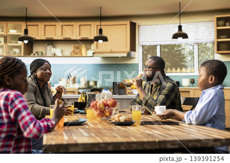 African American family sharing pastry and drinks for breakfast at home, parents serve scrambled eggs, toast and fruit. Cozy kitchen moment captures warmth, relaxation and joyful togetherness. African American family sharing pastry and drinks for breakfast at home, parents serve scrambled eggs, toast and fruit. Cozy kitchen moment captures warmth, relaxation and joyful togetherness. 139391254
